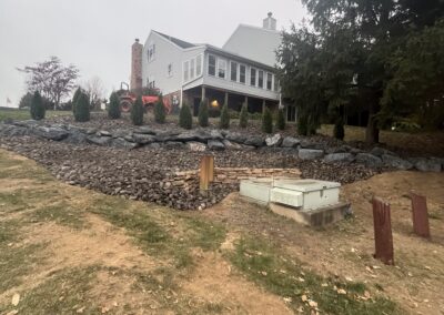 Two-story light-gray house on a sloped lot with a newly installed stacked boulder retaining wall and river rock slope, small evergreen shrubs planted along the wall, a tractor parked near the house, utility boxes and posts in the foreground, overcast sky