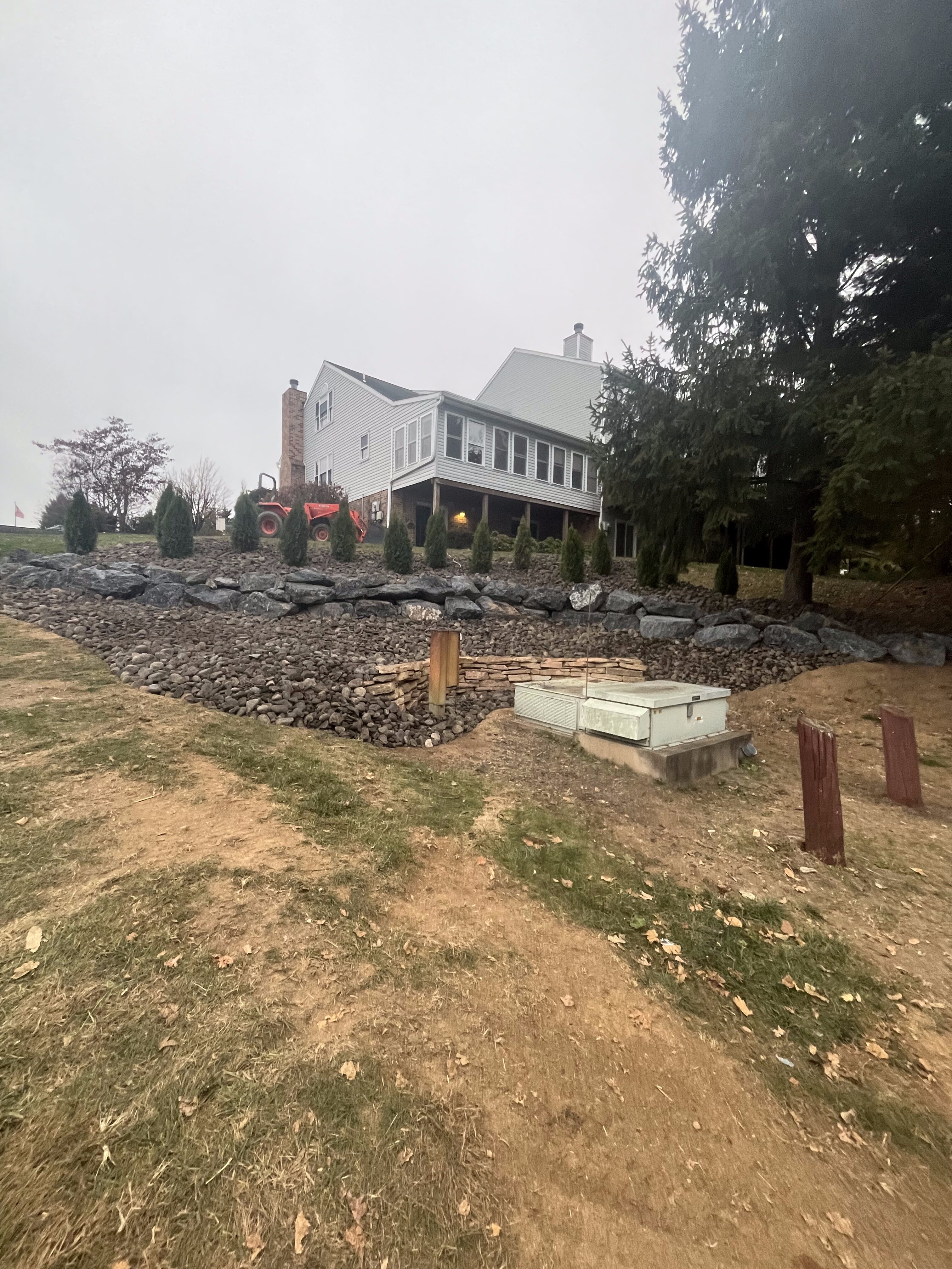 Two-story light-gray house on a sloped lot with a newly installed stacked boulder retaining wall and river rock slope, small evergreen shrubs planted along the wall, a tractor parked near the house, utility boxes and posts in the foreground, overcast sky