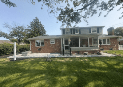 Rear view of a two-story brick and siding suburban house with a covered porch, stone patio, hanging planter, lawn and trees under a clear sky