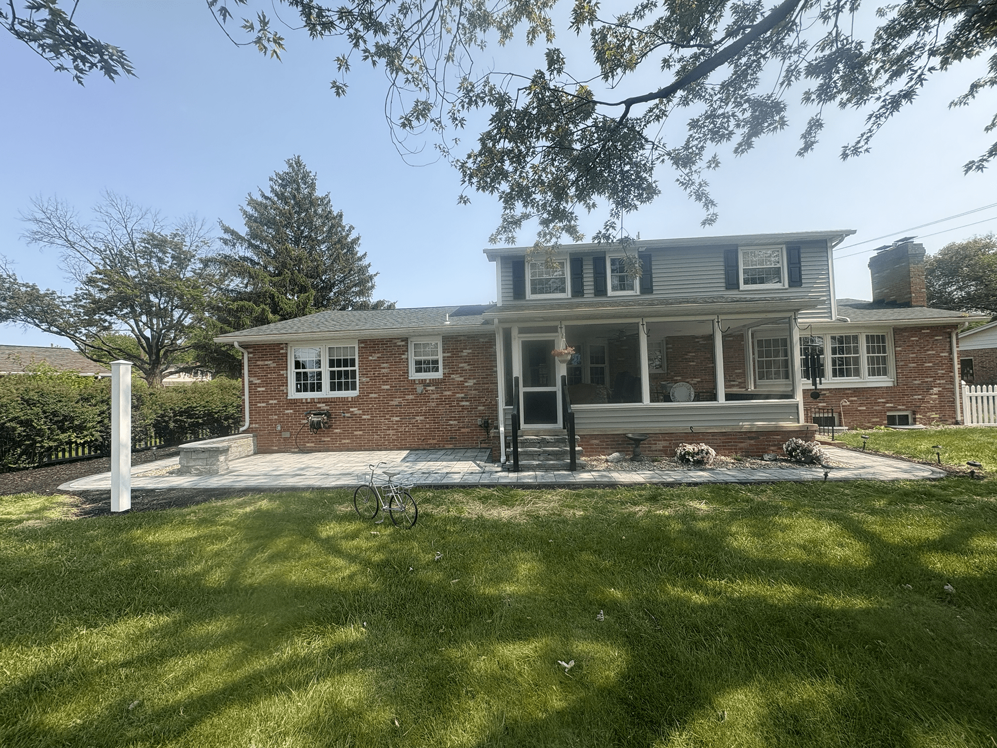 Rear view of a two-story brick and siding suburban house with a covered porch, stone patio, hanging planter, lawn and trees under a clear sky