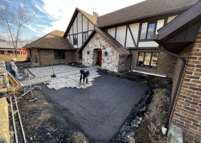 Worker laying large square pavers to form a new front patio/driveway area at a Tudor-style brick and stone house, with fresh compacted gravel/asphalt base, tools and materials around, bare trees and blue sky in background