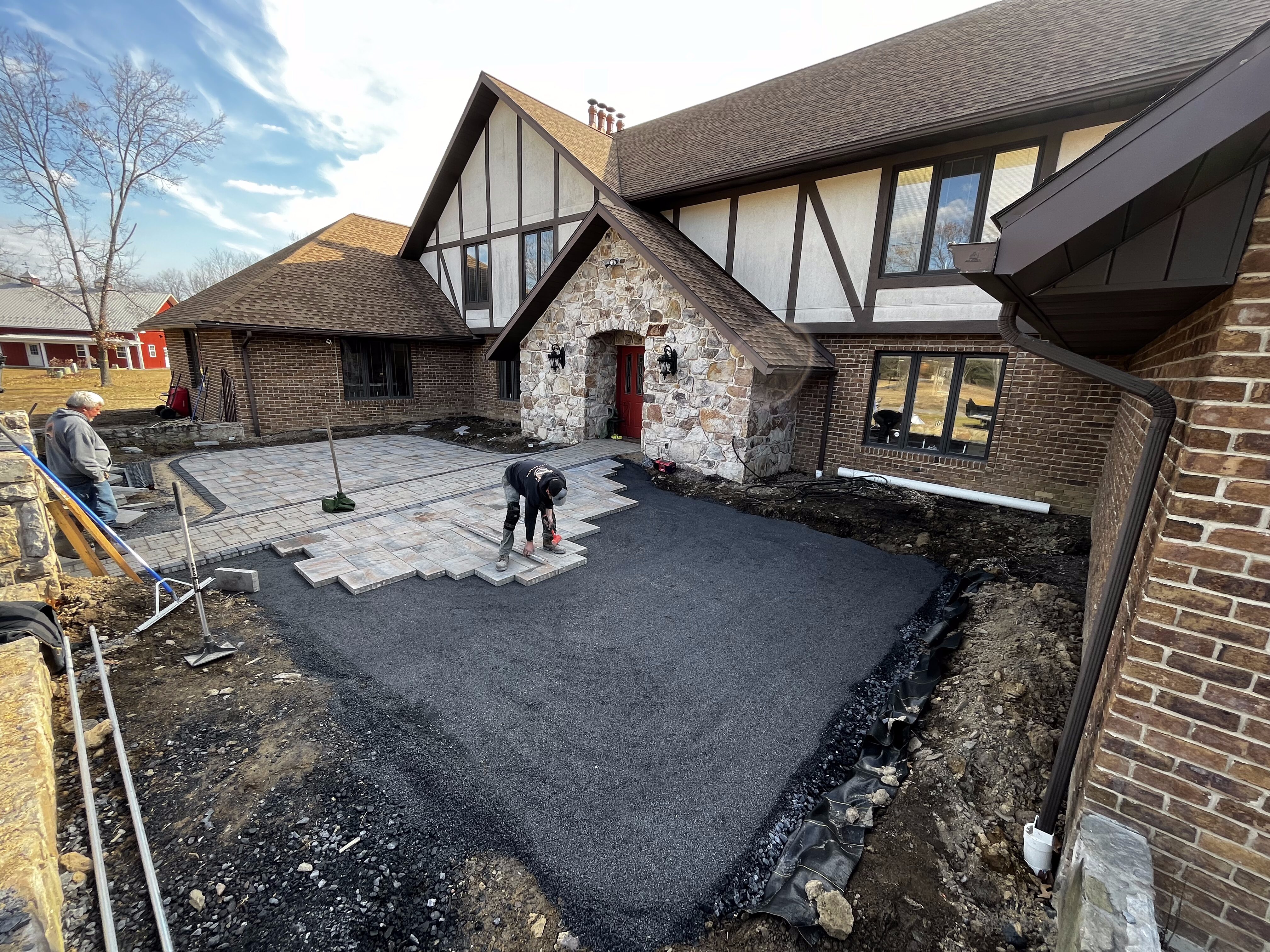 Worker laying large square pavers to form a new front patio/driveway area at a Tudor-style brick and stone house, with fresh compacted gravel/asphalt base, tools and materials around, bare trees and blue sky in background