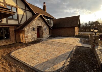 Stone-pattern stamped concrete driveway and walkway leading to a Tudor-style brick and stone house with a red front door, lantern-style wall lights, low stone border edging and cloudy late-afternoon sky