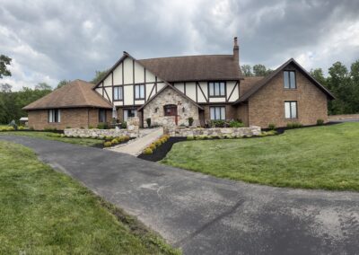 Large Tudor-style brick house with stone entryway and newly mulched planting beds and neatly mowed lawn, curved asphalt driveway leading to front walkway under a cloudy sky